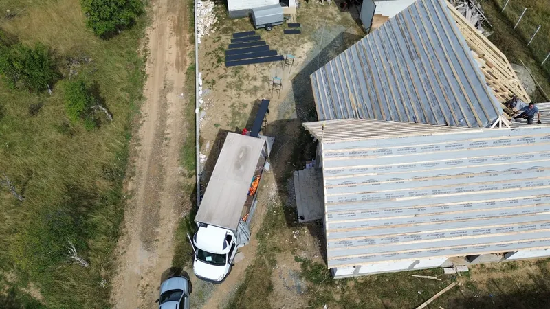 Vedere de sus: camionul cu linia de profilare parcat lângă casă, cu panouri profilate întinse în curte.
