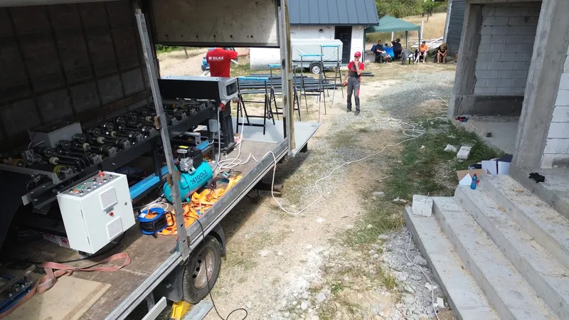 Utilajul de profilare în camionul deschis, lângă o casă în construcție; în curte, capre pentru panouri.
